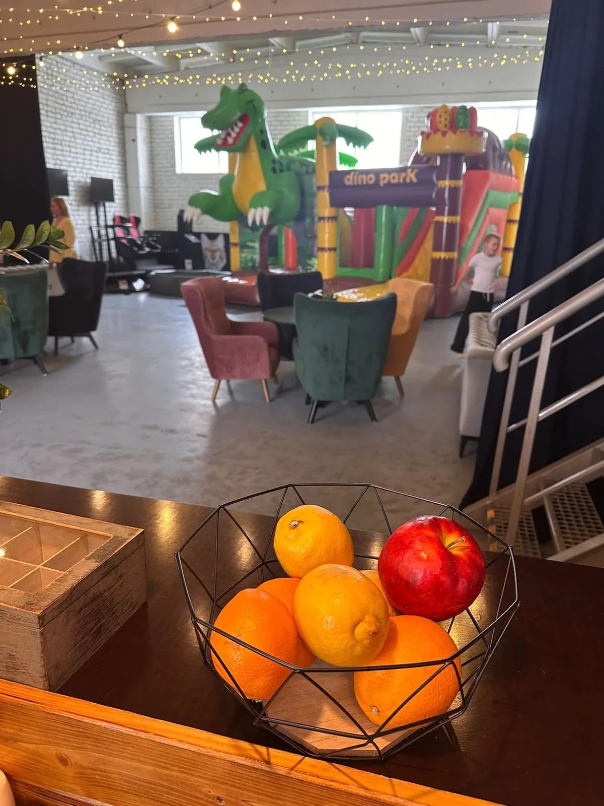 Kureti Cafe zāle: augļu groziņš uz bāra letes, aizmugurē samta krēsli, gaismas virtenes un piepūšamā atrakcija bērniem